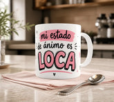 Taza de cerámica blanca con ilustración de una chica expresiva con el pelo alborotado y la frase humorística "mi estado de ánimo es LOCA".
